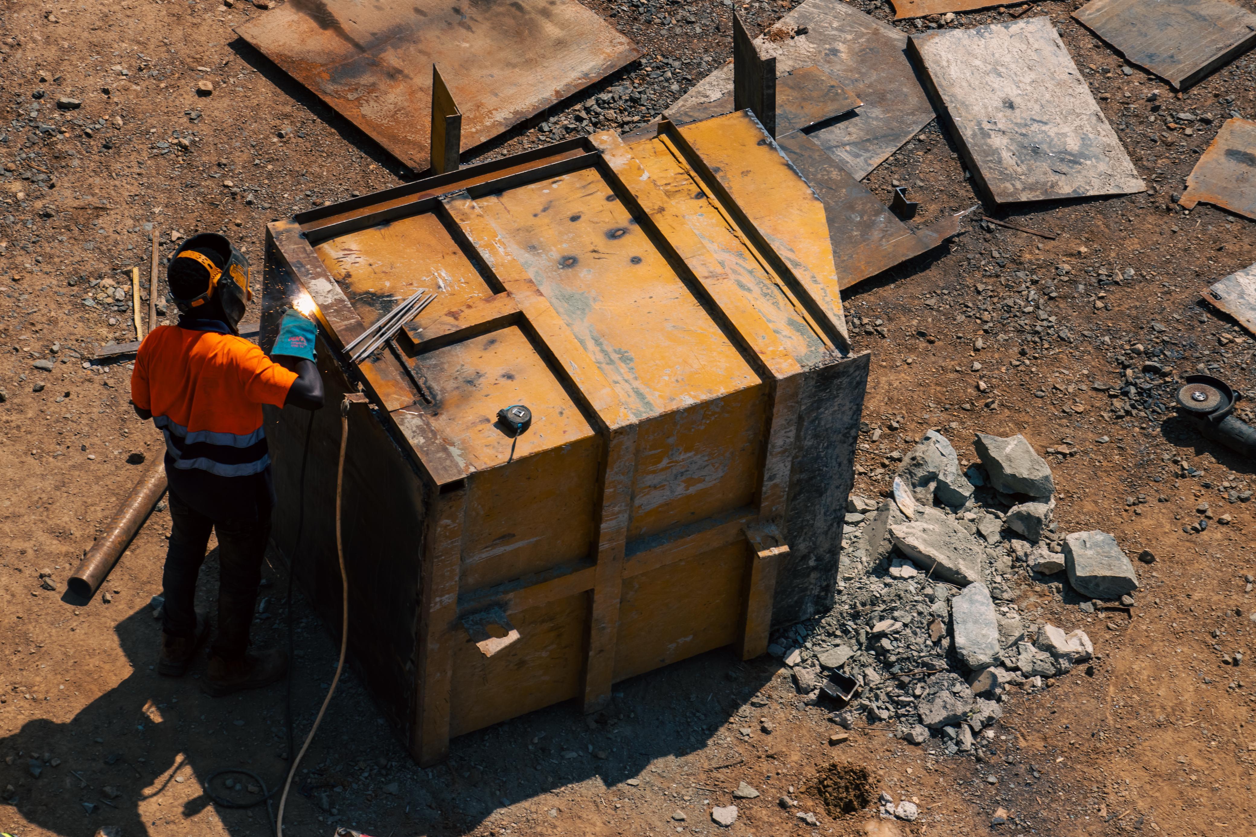 A worker welding a sealed steel transport crate at a mine site, seen from above.