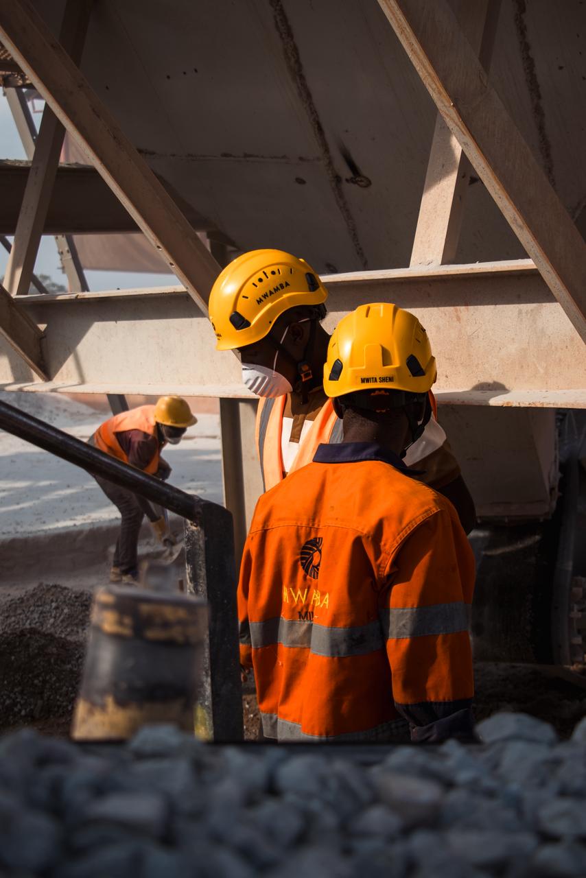 Workers in Mwamba Mining overalls at a gold processing plant, seen from behind.
